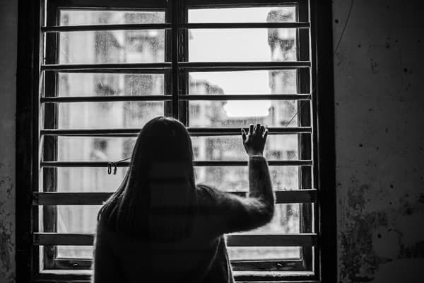 鉄格子の窓の前に立ち、外を見つめる長髪の女性。閉じ込められたような空気の中に、外の光が差し込んでいる様子。